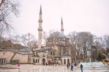 Amasya 'da görkemli bir cami var. Açık renkli taşları, etrafını saran çıplak kış ağaçlarıyla çelişiyor. Birkaç kişi meydanda yürür, merkez çeşmeye doğru. Bulutlu gökyüzü sahneye barışçıl bir atmosfer ekler..