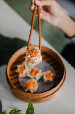 Female hand taking Chinese Dimsum with chopsticks in traditional bamboo basket on wooden table and green leaves background