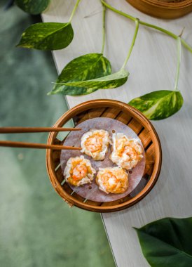 Female hand taking Chinese Dimsum with chopsticks in traditional bamboo basket on wooden table and green leaves background