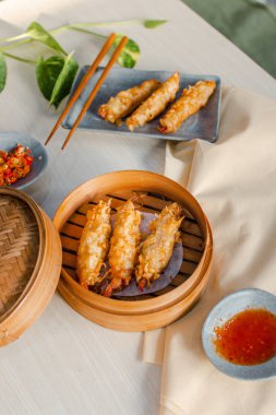 Fried tempura prawn Dimsum in traditional bamboo basket with tomato sauce, slice of chili on bowl and chopsticks on wooden background