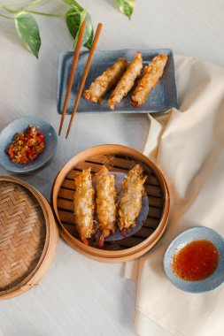 Fried tempura prawn Dimsum in traditional bamboo basket with tomato sauce, slice of chili on bowl and chopsticks on wooden background