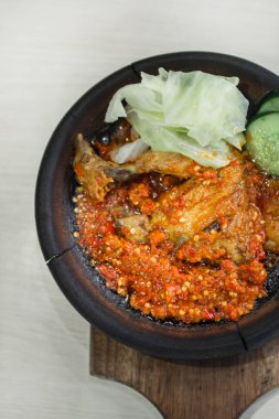 Close up fried chicken with sambal cooked in earthenware plate against wooden background. Served on wooden table with chili, spoon and fork as decoration
