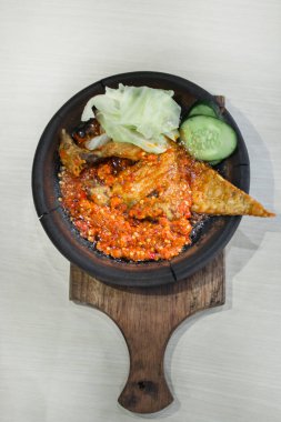 Close up fried chicken with sambal cooked in earthenware plate against wooden background. Served on wooden table with chili, spoon and fork as decoration