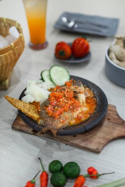 Close up sambal gami gurame or carp fish with sambal cooked in earthenware plate against wooden background. Served on wooden table with chili, spoon and fork as decoration
