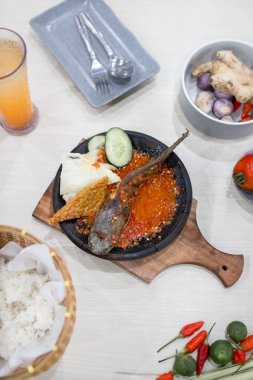 Close up gami lele or catfish with sambal cooked in earthenware plate against on wooden background. Serve on wooden table with chili, spoon and fork as decoration