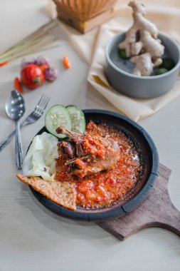 Sambal gami ayam goreng or fried chicken with sambal cooked in earthenware plate against wooden background. Served on wooden table