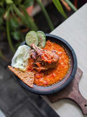 Sambal gami ayam goreng or fried chicken with sambal cooked in earthenware plate against wooden background. Served on wooden table