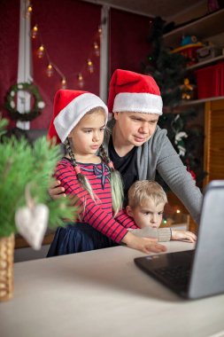 Güvenli çevrimiçi Noel kutlaması. Noel Baba kırmızı şapkalı mutlu aile arkadaşlarıyla internet ve defterle kutlama yapıyor. Görüntülü arama. Evde kal, uzak tatil, yaşam tarzı