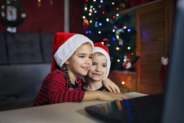 Safe online Christmas celebration. Happy kids in Santa red hats with burning sparkler celebrating with friends virtually via internet and notebook. Video call. Stay home, distant holiday lifestyle