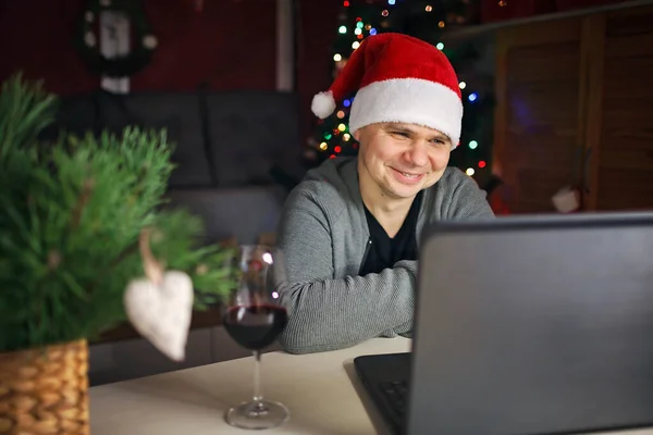 Online Christmas celebration. Happy man in Santa red hat celebrating with family virtually via internet and notebook. Video call. Refugees and immigrants, distant holiday, lifestyle