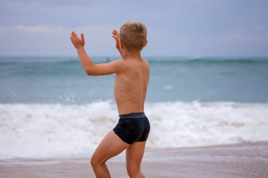 A boy of 7-8 years old sits on the ocean shore with his arms open towards the wind and waves. Storm on the ocean in summer. The concept of freedom, courage and challenge. Place for text