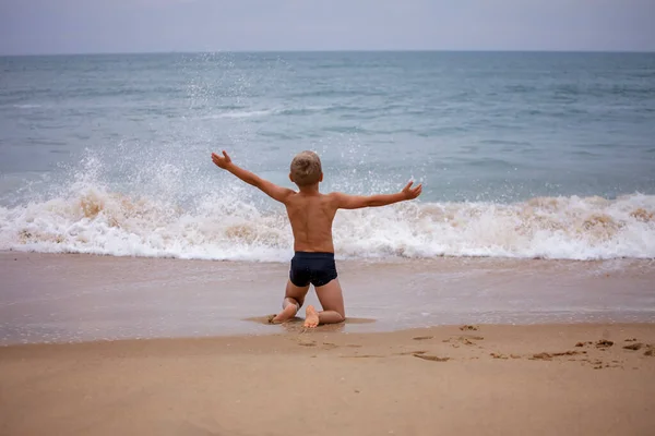 A boy of 7-8 years old sits on the ocean shore with his arms open towards the wind and waves. Storm on the ocean in summer. The concept of freedom, courage and challenge. Place for text