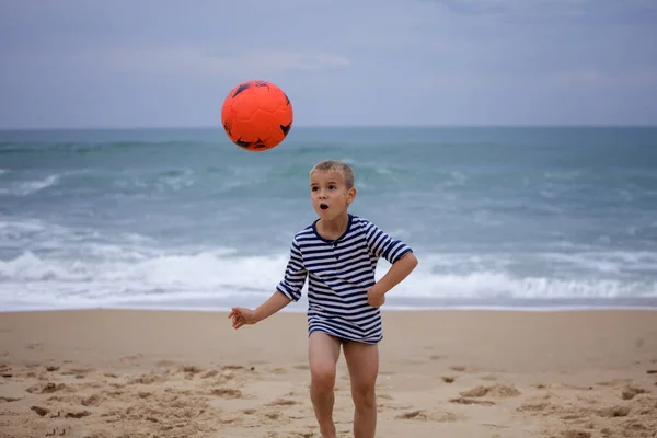 A small football fan barefoot plays football just on the band of ocean. Football everywhere. Sport, soccer, fandom concept. Storm outside and storm of emotions inside