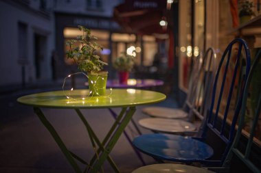 Alacakaranlıkta konforlu bir Paris kafe terası, saksı bitkisi barındıran yeşil bir masa ve parıldayan ışıklar, akşam rahatlığı ve sosyalleşme için davetkar bir atmosfer sunuyor. Seyahat ve seyahat kavramları