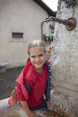 Kırmızı tişörtlü, gülümseyen genç bir kız, eski bir köy çeşmesinden akan suyla oynar. Güneşli bir günde basit, neşeli bir anın tadını çıkarır..