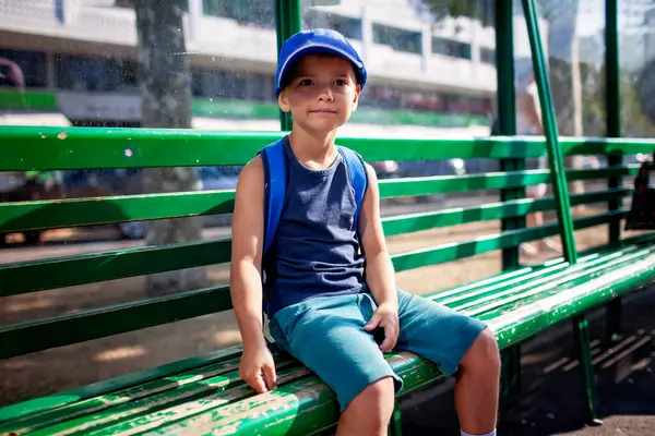 Genç çocuk bir otobüs durağında sabırla toplu taşıma bekliyor. Yazın şehir rutini, bağımsızlık, beklenti ve günlük yaşam sahneleri.