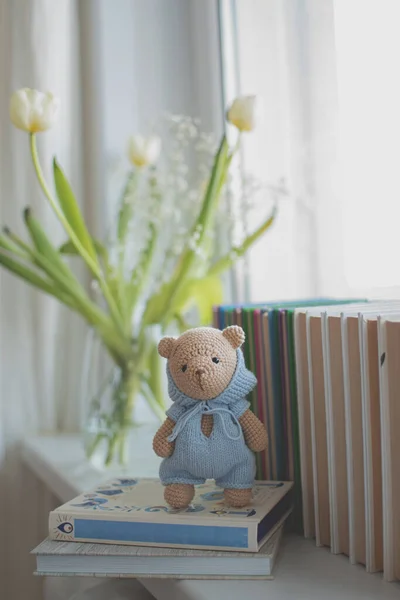 handmade knitted teddy bear in a blue jumpsuit stands on books, a vase with flowers