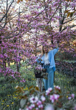 Güzel sarışın kız elinde bisikletle çiçek açan bahar bahçesinde duruyor.