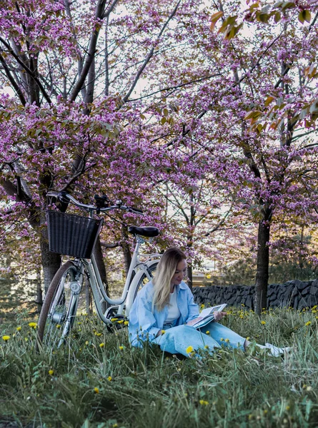 beautiful young woman sits in the grass reading a book under a tree with pink flowers