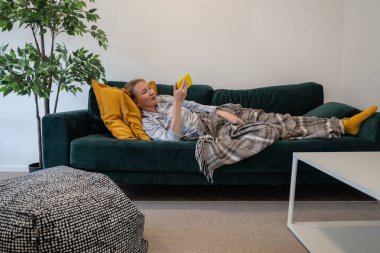 Pijamalı ve battaniyeli genç bir kadın kanepeye uzanıyor ve akıllı telefon kullanıyor.