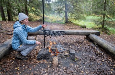 Mavi ceketli kadın ormanda kamp ateşinde yemek pişiriyor.