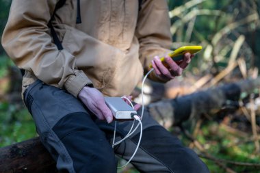 Adam ormanda elinde taşınabilir şarj cihazıyla akıllı telefonu şarj ediyor.
