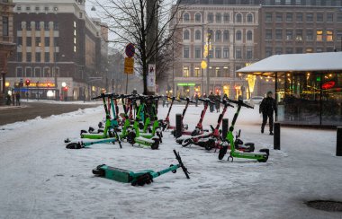 Helsinki, Finlandiya - 23 Kasım 2024: Karla kaplı elektrikli scooterlar karlı bir caddeye park edilmiş, arka planda kentsel binalar ve ağaçlar kentsel taşımacılığı simgeliyor