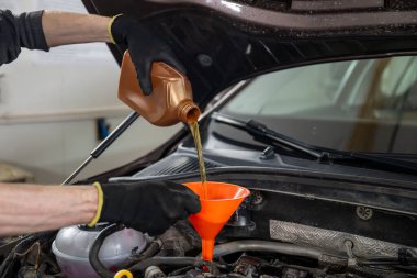 Mechanic in gloves pouring motor oil into a car engine using an orange funnel, in a workshop. Concept of vehicle maintenance clipart