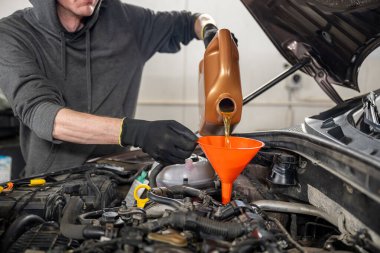 Mechanic in gloves pouring motor oil into a car engine using an orange funnel, in a workshop. Concept of vehicle maintenance clipart