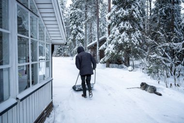 Kırsal ahşap bir kulübede kar temizleyen bir adam. Kış ormanıyla çevrili.