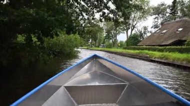 Hollanda, Giethoorn 'da yemyeşil ağaçlar ve çimlerle çevrili dar bir kanalda yüzen metal tekne.