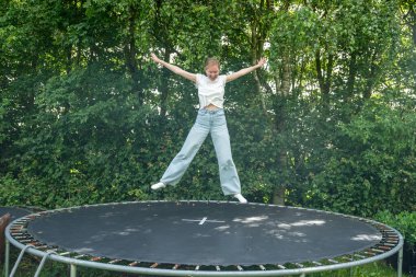 Kadın trambolinde kot pantolon ve yeşil ormanın yanındaki beyaz gömlekle zıplıyor.