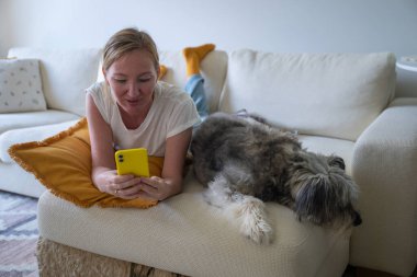 Kadın rahat oturma odasında akıllı telefon kullanırken köpekle kanepeye uzanmış..