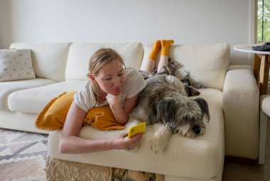 Kadın rahat oturma odasında akıllı telefon kullanırken köpekle kanepeye uzanmış..