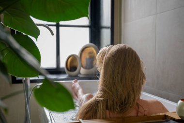 Pencerenin yanındaki küvette dinlenen bir kadın. Etrafında çiçekler ve mumlarla..