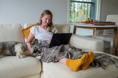 Parlak çoraplı kadın krem rengi koltukta oturuyor, battaniyesi ve köpeği var ve dizüstü bilgisayar kullanıyor..