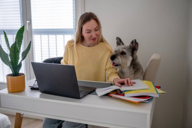 Sarı kazaklı bir kadın evde köpeğiyle birlikte dizüstü bilgisayarda çalışıyor. Uzaktan çalışma kavramı. 