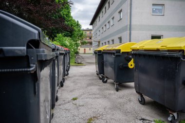 Konut binalarının yakınında siyah, kırmızı ve sarı kapaklı çok sayıda çöp konteynırı var. . 