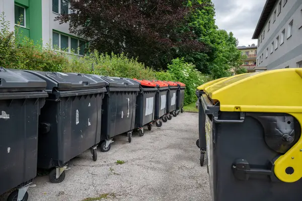 Konut binalarının yakınında siyah, kırmızı ve sarı kapaklı çok sayıda çöp konteynırı var. . 