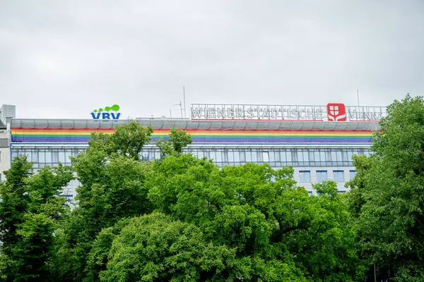 Viyana, Avusturya - 30 Mayıs 2025: Gökkuşağı sancağı ve bulutlu gökyüzü altındaki ağaçlarla modern ofis binası. 