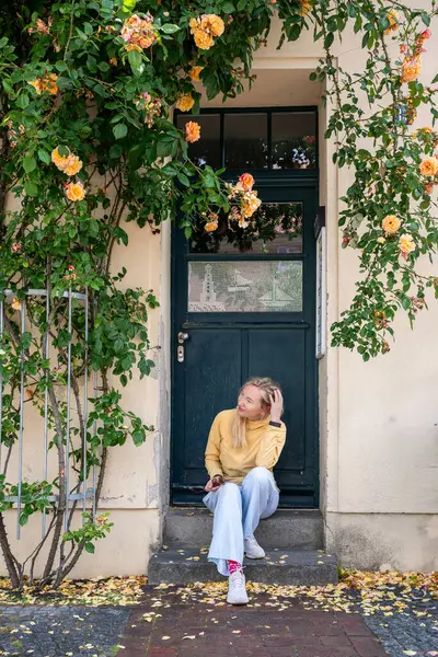 Genç bir kadın, çiçeklenmiş gül çalılarının altında, eski evlerin önünde oturuyor.. 