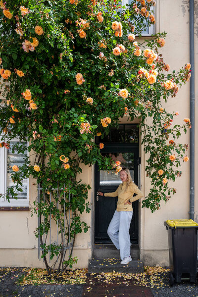 Молодая женщина сидит на пороге под цветущим розовым кустом перед винтажным фасадом дома. 