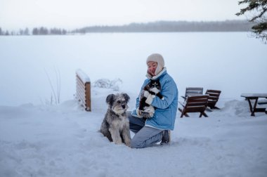 Mavi kışlık ceketli bir kadın, karlı gölün kenarında kedisi ve tüylü bir köpeği var..