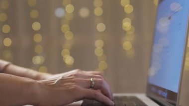 Bir kadın Noel tatilinde çalışır. Bir kız, yılbaşı ışıkları yanıp sönen bir fotoğrafın önünde dizüstü bilgisayarda yazı yazıyor. Yakın çekim video. Yüksek kalite 4k görüntü