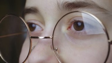 A teenage girl in round glasses with a gold rim looks into space. The teenager adjusts his glasses with his finger, which have slipped down his nose. Cute girl with brown eyes wearing glasses. High