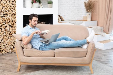 Yakışıklı adam kanepede uzanıyor, kitap okuyor ve dizüstü bilgisayarla çalışıyor. Çevrimiçi eğitim ve mesafe çalışma kavramı