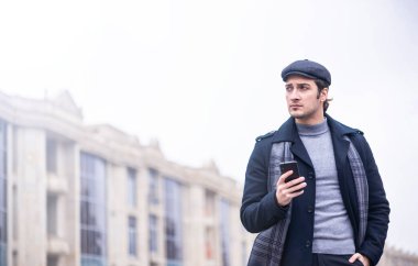 Siyah ceketli yakışıklı bir erkek model ve açık havada duran atkısı olan gri düz bir şapka.