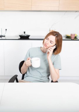 Ciddi bir kadın mutfakta oturmuş elinde bir fincan kahveyle telefonla konuşuyor.