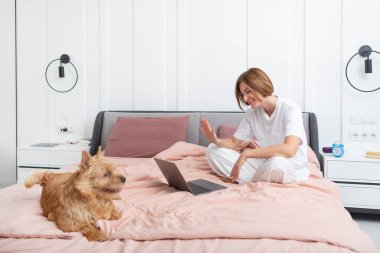 Yatakta oturan sevimli köpek portresi ve yatak odasında arka planda yatan ve gülümseyen kadın.