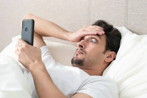 令人惊讶和震惊的男人躺在床上 早上看着智能手机 在坏消息的概念和检查手机后醒来 — 图库照片